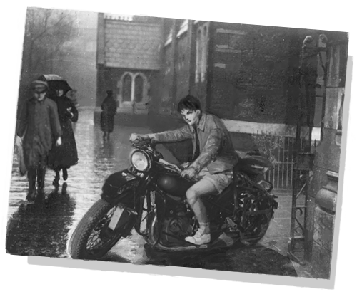 a black and white photo of a rainy day. the cobbled street is wet, and people are walking quickly by. a brick building sits in the background and disappears to the right. in the foreground is Levi on a motercycle. he is wearing a collared coat and shorts. in this weather? smh